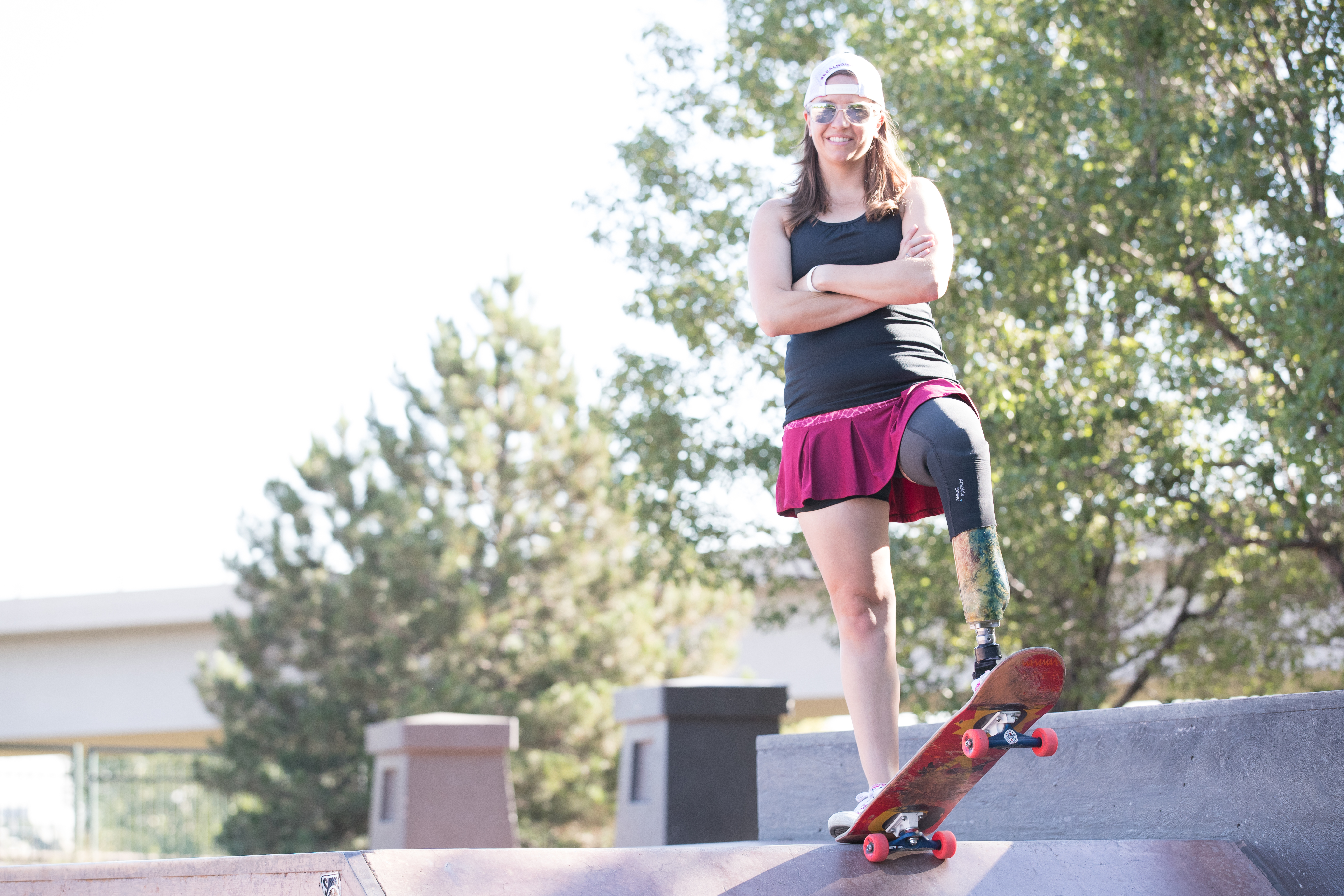 Emily at the skate park for a Skirt Sports photo shoot. Wearing a pink skirt, black top, backwards trucker hat, and sunglasses. One foot on the back of a skateboard and the other resting on the front, arms crossed