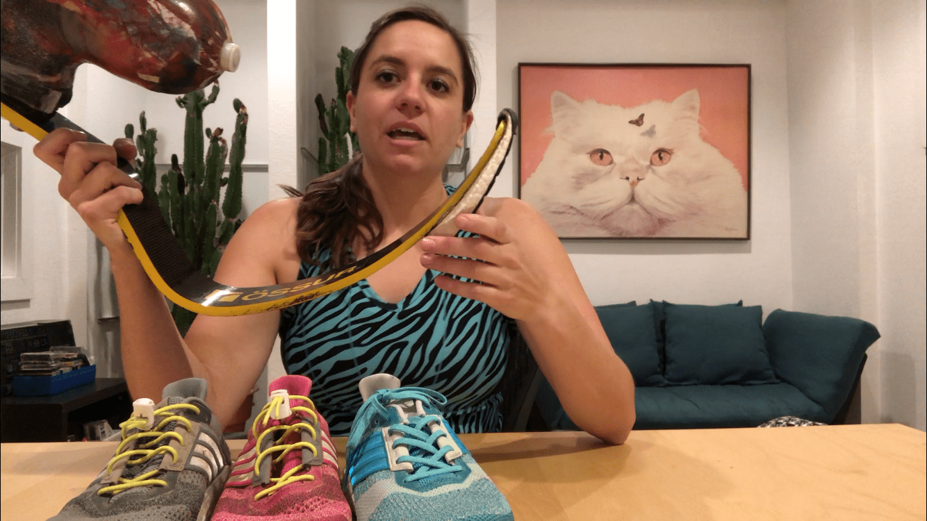 Screen shot of video where Emily talks about running shoes - she is holding up her running leg and there are 3 running shoes of different colors also in the shot