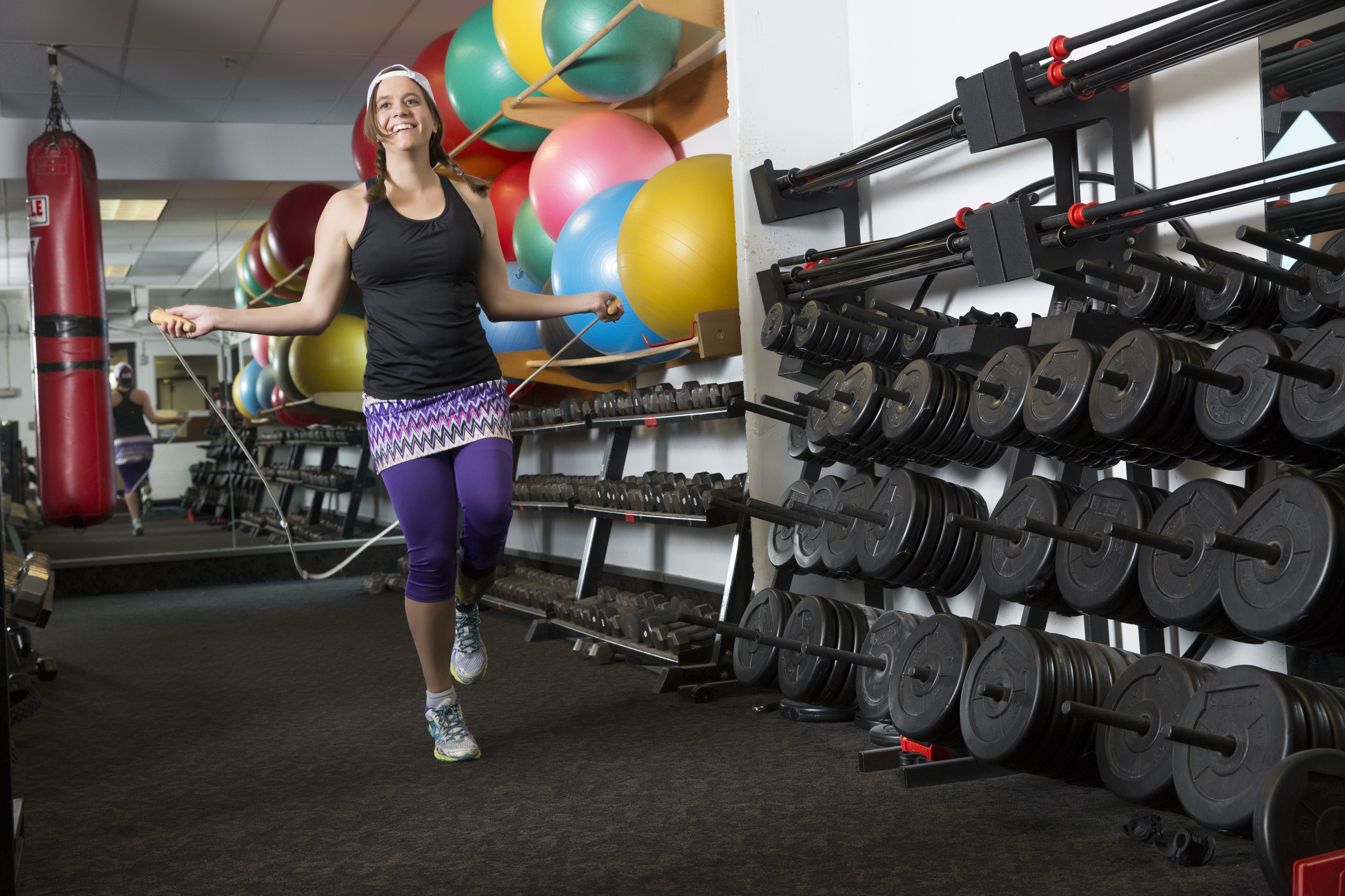 A woman with brown hair and a prosthetic leg is jumping rope in a gym. She is wearing colorful athletic clothes.